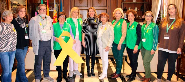 Begoña Carrasco recibe de manos de la gerente de la Asociación Española contra el Cáncer de Castellón, Salomé Esteller, el lazo amarillo conmemorativo e insta a seguir aunando esfuerzos para avanzar en la investigación que salva vidas.